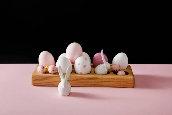 Coelhinhos brancos decorativos e ovos de páscoa em tábua de madeira sobre fundo rosa e preto — Fotografia de Stock
