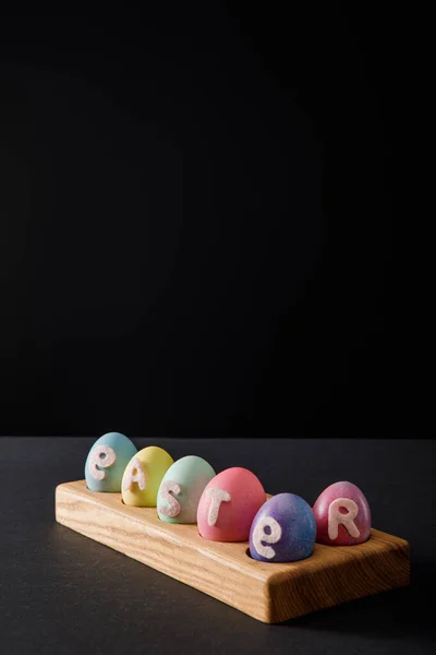 Painted chicken eggs with easter lettering on wooden board on grey and black background — Stock Photo