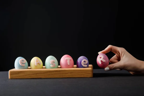 Vista recortada de la mujer sosteniendo huevo de gallina pintado cerca de la tabla con huevos de colores con letras de Pascua en gris y negro - foto de stock