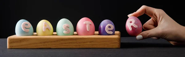 Vue recadrée de la femme tenant un œuf de poulet peint près du bord avec des œufs colorés avec des lettres de Pâques sur gris et noir, prise de vue panoramique — Photo de stock