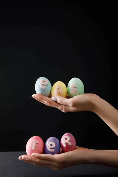 Vue recadrée de la femme tenant des œufs de poulet peints avec lettrage de Pâques sur fond noir — Photo de stock
