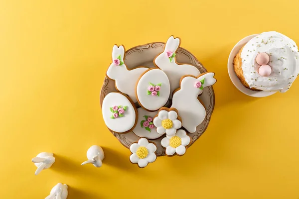 Vista superior da tigela com biscoitos saborosos, coelhos decorativos e bolo de Páscoa no fundo amarelo — Fotografia de Stock