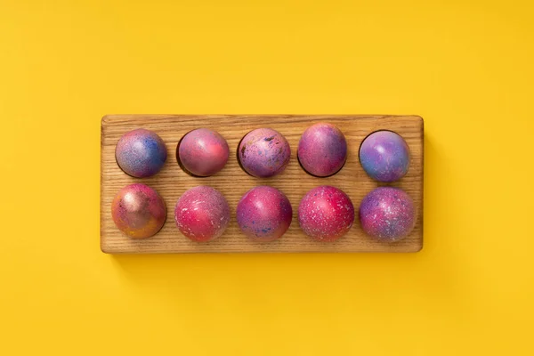 Vue du dessus de la planche en bois avec des œufs de Pâques colorés sur fond jaune — Photo de stock