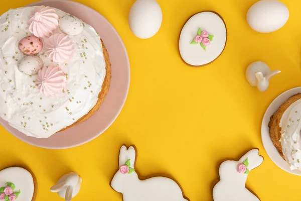 Vista superior de pasteles de Pascua con huevos de pollo, galletas y conejos decorativos sobre fondo amarillo - foto de stock