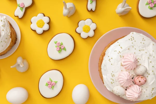 Vista superior de pasteles de Pascua, huevos de pollo, galletas y conejos decorativos sobre fondo amarillo - foto de stock