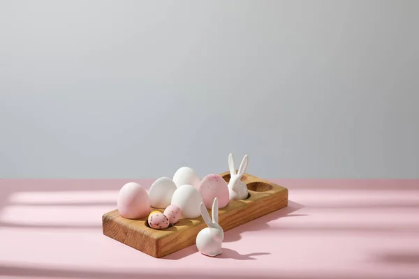 Easter eggs on wooden board with decorative rabbits on pink and grey background — Stock Photo