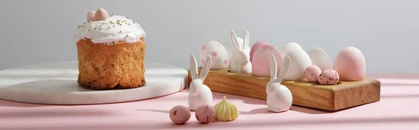 Пасхальні яйця на дерев'яній дошці з декоративними кроликами біля великоднього торта на рожевому та сірому фоні — стокове фото