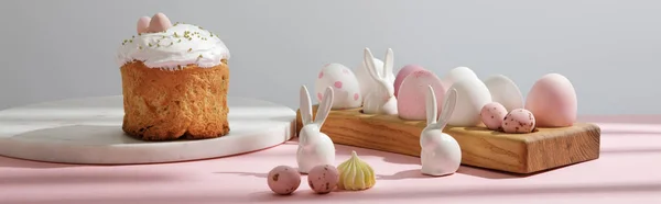 Easter eggs on wooden board with decorative rabbits near easter cake on pink and grey background — Stock Photo