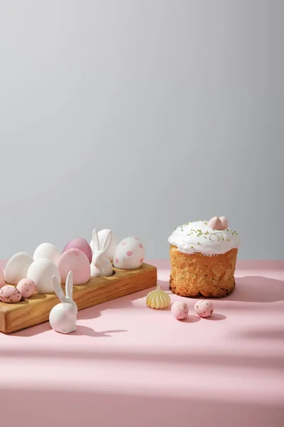 Uova di Pasqua su tavola di legno con conigli decorativi vicino torta pasquale su sfondo rosa e grigio — Foto stock