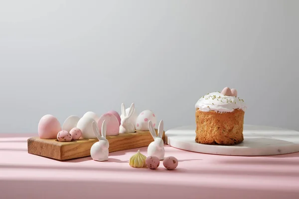 Ovos de Páscoa em tábua de madeira com coelhos decorativos perto de pão de Páscoa em fundo rosa e cinza — Fotografia de Stock
