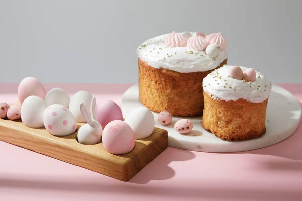 Великодні яйця на дерев'яній дошці з декоративним кроликом біля великодніх тортів на рожевому та сірому — стокове фото