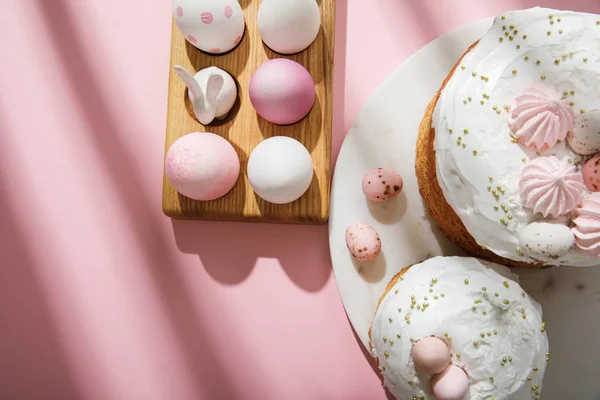 Top view of easter eggs on wooden board with decorative bunny near easter cakes on pink — Stockfoto