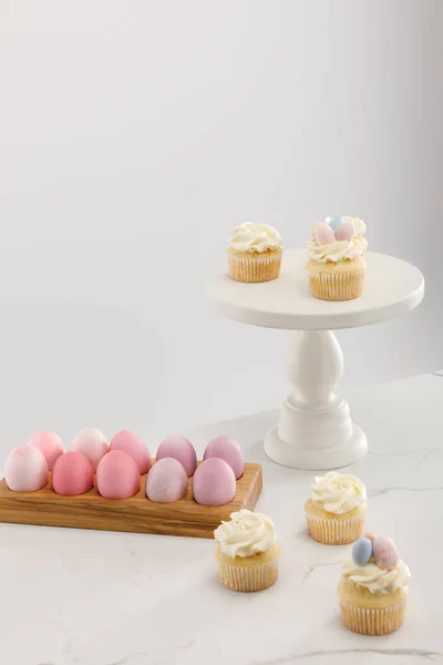 Delicious Easter cupcakes on surface and cake stand with painted chicken eggs on grey background — Stockfoto
