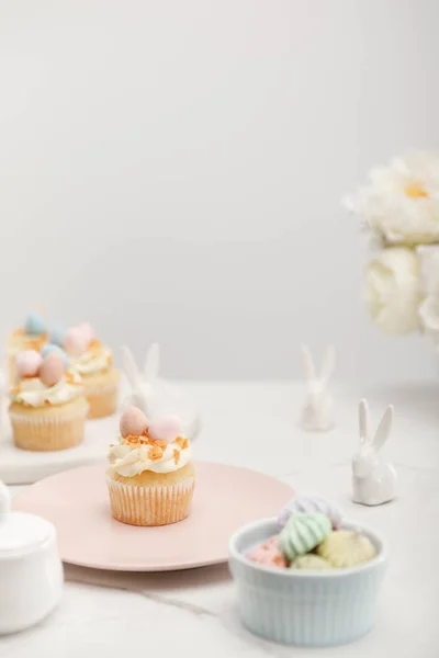 Enfoque selectivo de cupcakes con conejitos decorativos, bowl con merengues y flores sobre fondo gris - foto de stock