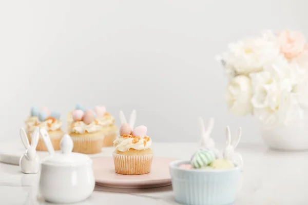 Foco seletivo de cupcakes, coelhos decorativos, açucareiro, merengues e vaso com flores em fundo cinza — Fotografia de Stock
