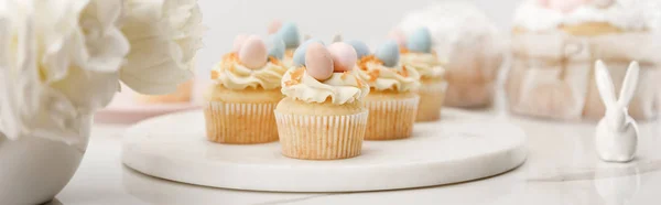 Foco seletivo de cupcakes na tábua redonda, coelho decorativo e vaso com flores no fundo branco, tiro panorâmico — Fotografia de Stock