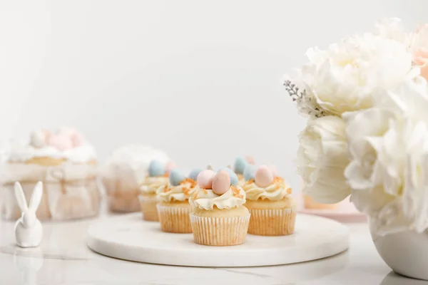 Enfoque selectivo de cupcakes sobre tabla redonda, conejito decorativo y jarrón con flores sobre fondo blanco - foto de stock