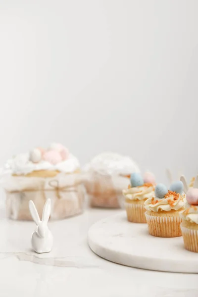 Селективное внимание кексы на круглом столе, декоративные кролики и пасхальные торты на сером фоне — стоковое фото