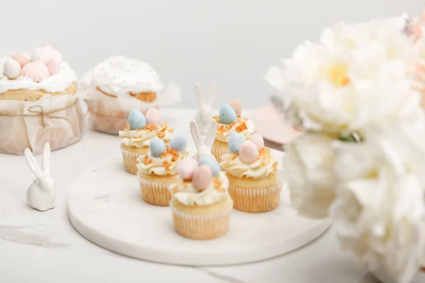 Selective focus of cupcakes with decorative bunnies on round board, flowers and easter cakes isolated on grey — Stockfoto