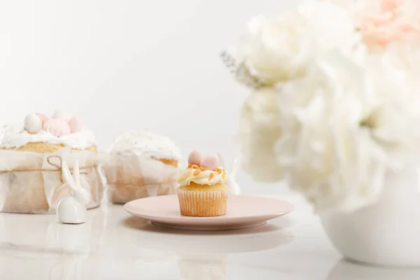 Foco seletivo de cupcake na placa, coelhos decorativos, bolos de Páscoa e vaso com buquê no fundo branco — Fotografia de Stock