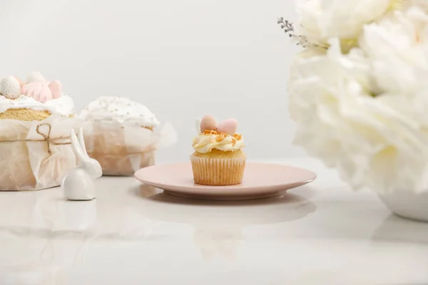 Foco seletivo de cupcake na placa, coelhos decorativos perto do vaso com buquê e bolos de Páscoa isolados em cinza — Fotografia de Stock
