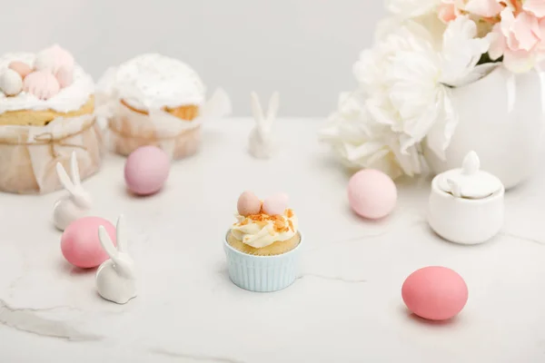 Foco seletivo de cupcake com ovos de galinha coloridos, coelhos decorativos, tigela de açúcar, bolos de Páscoa e flores isoladas em cinza — Fotografia de Stock