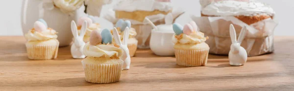 Enfoque selectivo de cupcakes con conejitos decorativos, azucarero, pasteles de Pascua y jarrón sobre fondo de madera, plano panorámico - foto de stock