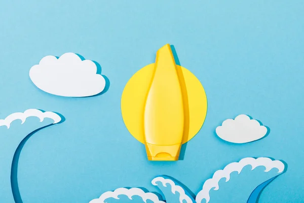 Vue du dessus du tube de crème solaire avec du papier coupé soleil, nuages et vagues de mer sur fond bleu — Photo de stock