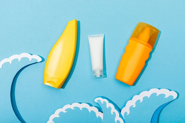 Top view of tubes and dispenser bottle of sunscreen with paper cut sea waves on blue background — Stock Photo