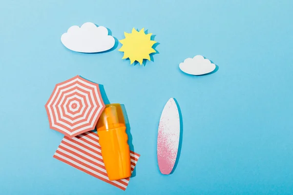Vue de dessus de la plage de papier découpé avec planches de surf, parapluie, couverture et bouteille de crème solaire sur bleu — Photo de stock