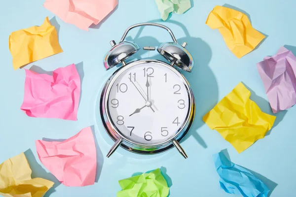 Vista superior del reloj despertador clásico con notas adhesivas coloridas arrugadas sobre fondo azul - foto de stock