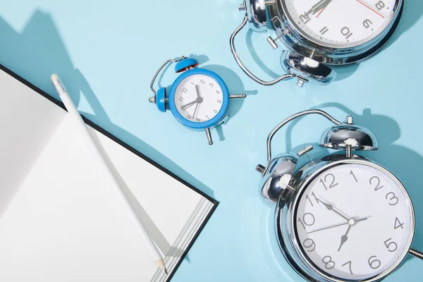 Vista superior de los relojes de alarma clásicos con netbook en blanco y lápiz sobre fondo azul - foto de stock