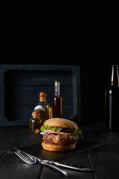 Foco selectivo de hamburguesa en la tabla de cortar negro cerca de cubiertos, aceite, vinagre y botellas de cerveza aislados en negro - foto de stock