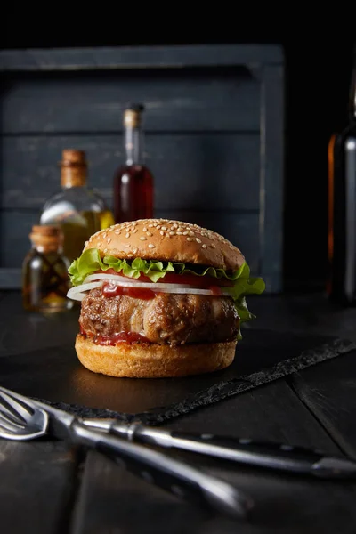 Foco seletivo de hambúrguer em placa de corte escuro perto de garfo, faca, óleo e vinagre garrafas isoladas em preto — Fotografia de Stock