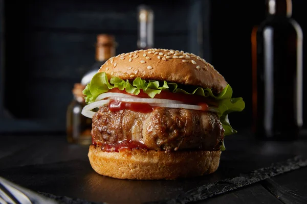 Selective focus of tasty burger on dark surface, beer, vinegar and oil bottles isolated on black — Stock Photo