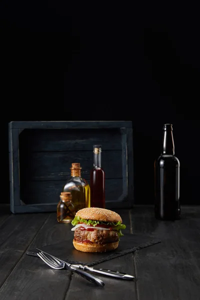 Hamburguesa en la superficie de madera oscura, tenedor, cuchillo y botellas de aceite aislados en negro - foto de stock