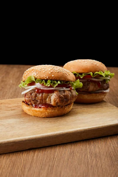 Two tasty burgers on wooden cutting board isolated on black — Stock Photo