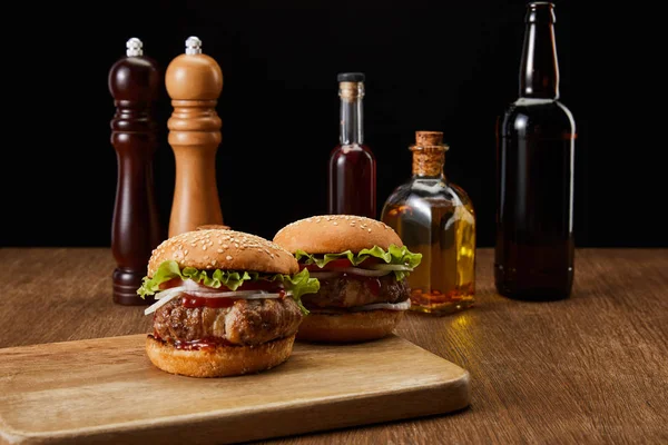 Two burgers on wooden chopping board, oil, beer and vinegar bottles, pepper and salt mills isolated on black — Stock Photo
