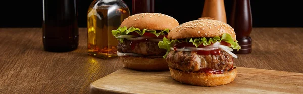 Panoramic shot of two tasty burgers on wooden board, cropped oil bottles, salt and pepper mills isolated on black — Stock Photo