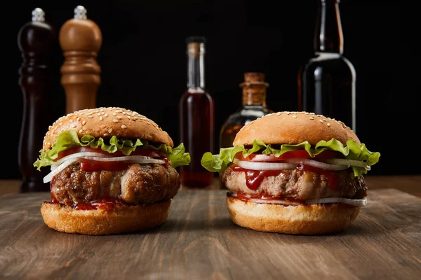 Selective focus of two burgers on wooden surface near oil, vinegar and beer bottles, pepper and salt mills isolated on black — Stock Photo