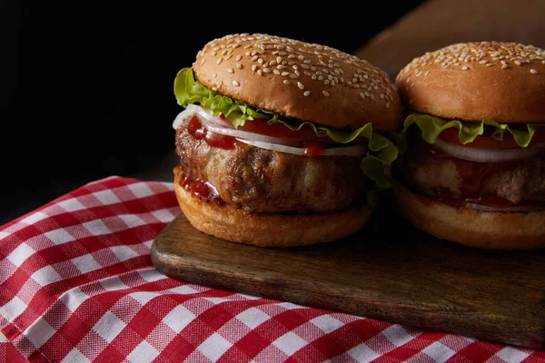 Two burgers on wooden cutting board on checkered tablecloth isolated on black — Stock Photo