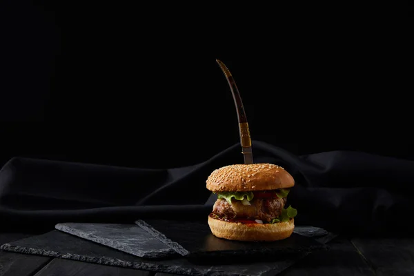 Delicious burger on black cutting boards pierced by knife isolated on black — Stock Photo