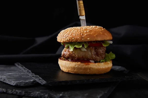 Burger pierced by knife on dark wooden cutting board isolated on black — Stock Photo