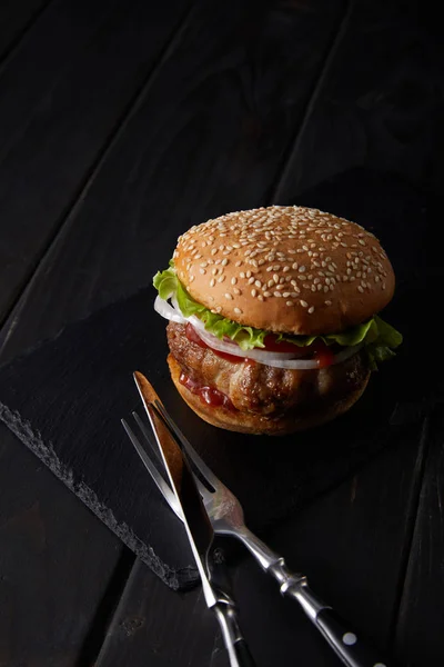 High angle view of a burger, fork and knife on a black wooden surface — Stock Photo