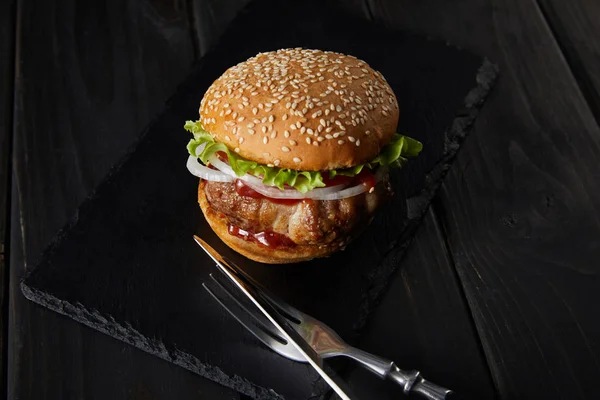 Burger on dark chopping board and cutlery on black wooden table — Stock Photo