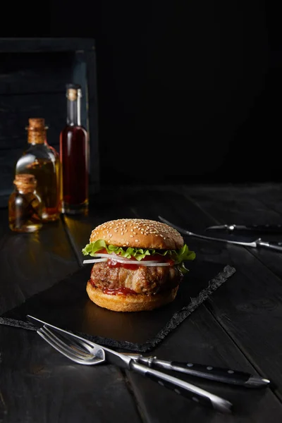 Foco selectivo de hamburguesa en la tabla de cortar oscura, tenedor, cuchillo, aceite y vinagre botellas aisladas en negro - foto de stock