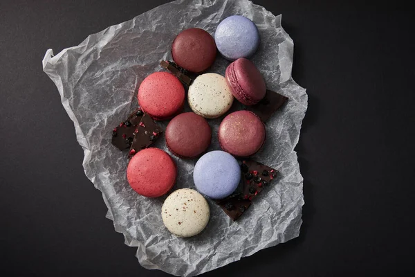Draufsicht auf verschiedene köstliche bunte französische Makronen auf zerknülltem Papier mit Schokolade auf schwarzem Hintergrund — Stockfoto