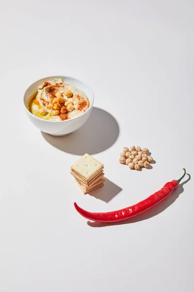 Cuenco con sabroso hummus cerca de galletas saladas, chile y garbanzo sobre fondo gris - foto de stock