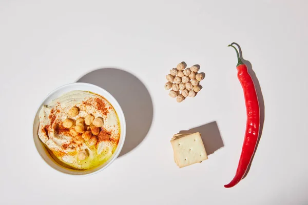 Top view of bowl with hummus near crackers, chili pepper and chickpea on grey — Stock Photo