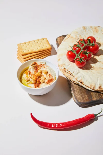 Bowl with hummus near crackers, chili and cutting board with pita bread and cherry tomatoes on grey — Stock Photo